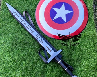 May include: A silver sword with a black leather scabbard and a red gem on the pommel. The sword has the words "I AM THE" engraved on the blade. The sword is lying on a green surface next to a red, white, and blue shield with a white star in the center.