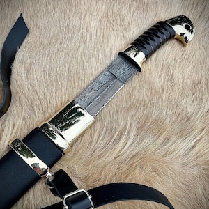 May include: A black leather scabbard with a gold-trimmed handle and a black and silver patterned blade. The scabbard has a black leather strap with a metal buckle.