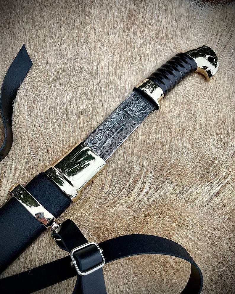 May include: A black leather scabbard with a gold-trimmed handle and a black and silver patterned blade. The scabbard has a black leather strap with a metal buckle.