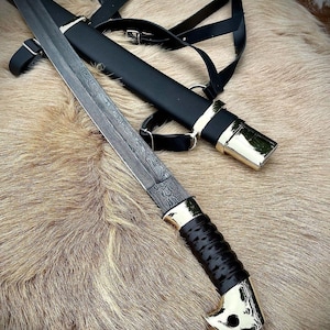 Könnte beinhalten: Ein schwarzes Lederscheide mit einer goldfarbenen Metallspitze und einem schwarzen Lederriemen. Eine silberfarbene Klinge mit einem schwarzen Griff ist teilweise sichtbar.