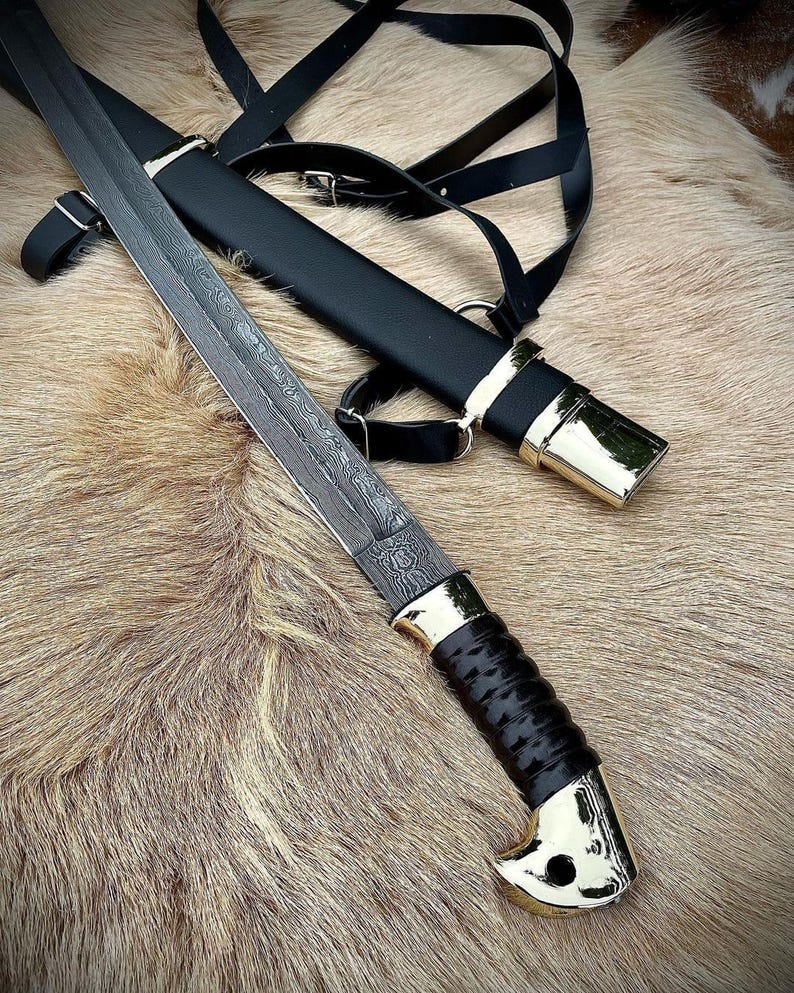 May include: A black leather scabbard with a gold-coloured metal tip and a black leather strap. A silver-coloured blade with a black handle is partially visible.