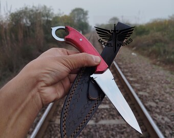 May include: A red-handled knife with a silver blade is held in front of a black leather sheath. The knife has a ring at the end of the handle. The sheath is brown stitched. The text "MODEL KNIVES" is visible.