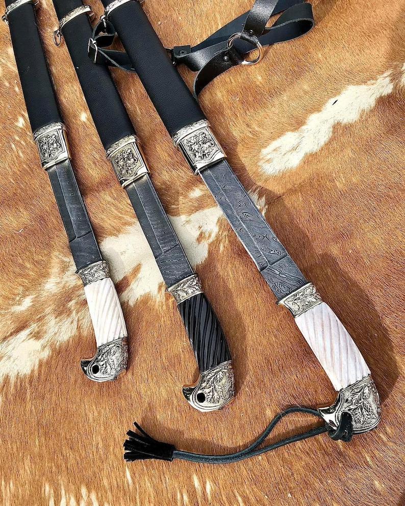 K&ouml;nnte beinhalten: Drei verzierte Schwerter mit schwarzen und wei&szlig;en Griffen und aufwendigen Metallarbeiten auf einem braunen und wei&szlig;en Pelz-Hintergrund.