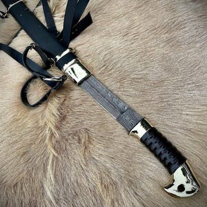 May include: A black leather scabbard with a silver-coloured metal tip and a black handle. The scabbard is attached to a brown leather strap with a silver-coloured metal buckle. The scabbard is holding a silver-coloured metal knife with a black handle.