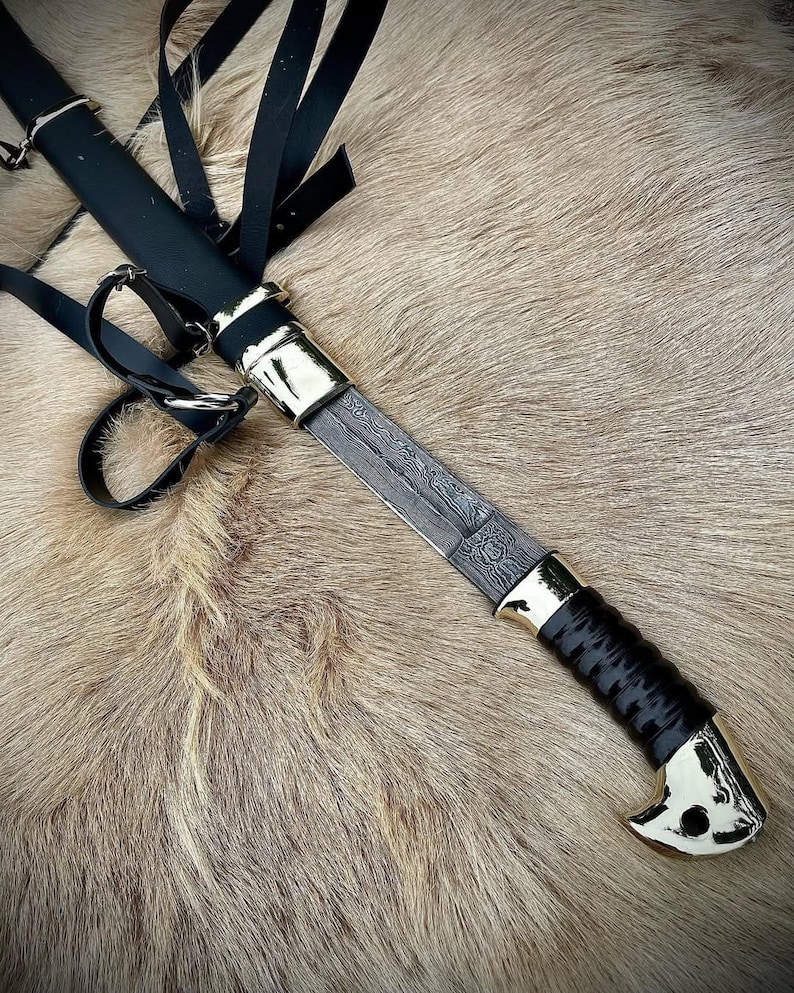 May include: A black leather scabbard with a silver-coloured metal tip and a black handle. The scabbard is attached to a brown leather strap with a silver-coloured metal buckle. The scabbard is holding a silver-coloured metal knife with a black handle.