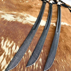 K&ouml;nnte beinhalten: Drei schwarze Schwerter mit komplizierten Mustern auf den Klingen. Die Schwerter liegen auf einer braunen Fellfl&auml;che.