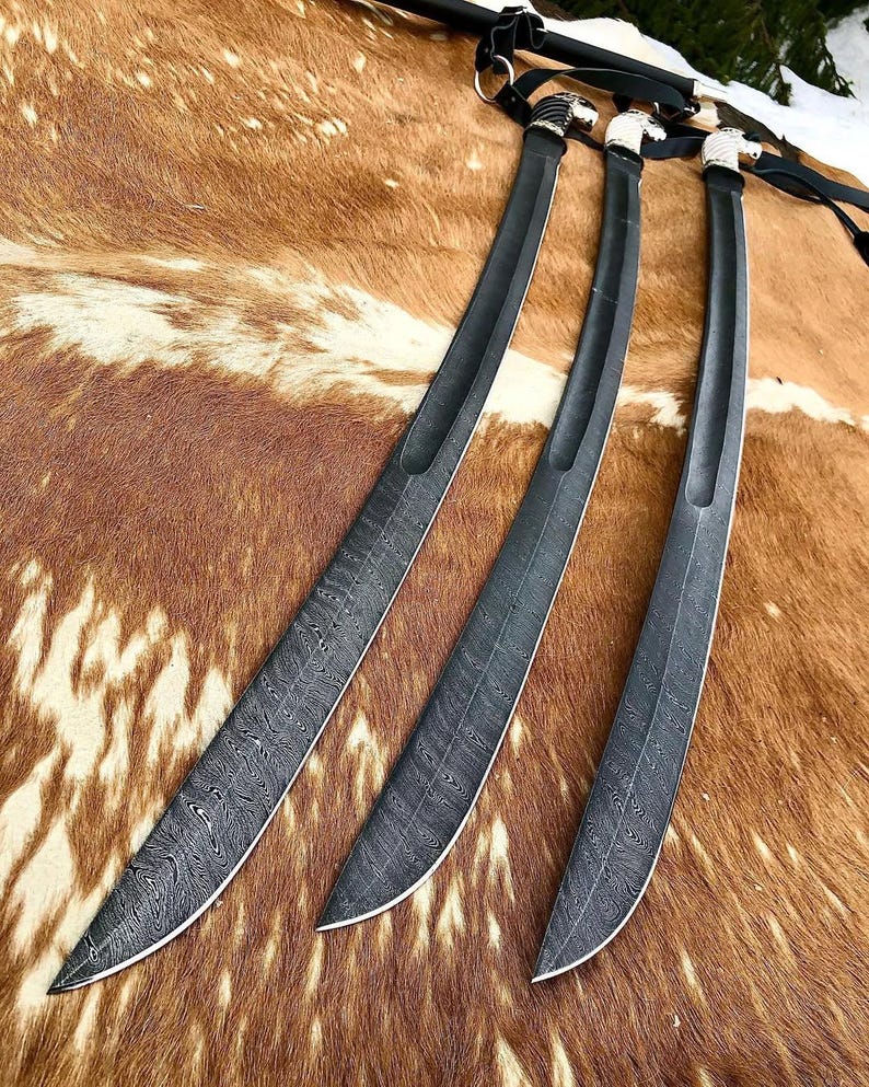 K&ouml;nnte beinhalten: Drei schwarze Schwerter mit komplizierten Mustern auf den Klingen. Die Schwerter liegen auf einer braunen Fellfl&auml;che.