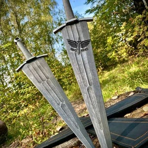May include: Two silver swords with intricate patterns on the blades. The swords are resting on a black leather sheath with orange stitching. The swords have a silver finish and are made of Damascus steel. The swords are displayed against a backdrop of green trees.