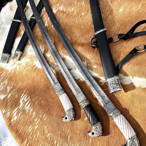 Könnte beinhalten: Drei verzierte Schwerter mit aufwendigen Metallarbeiten und weißen und schwarzen Griffen. Die Schwerter liegen auf einer braunen und weißen Tierhaut.