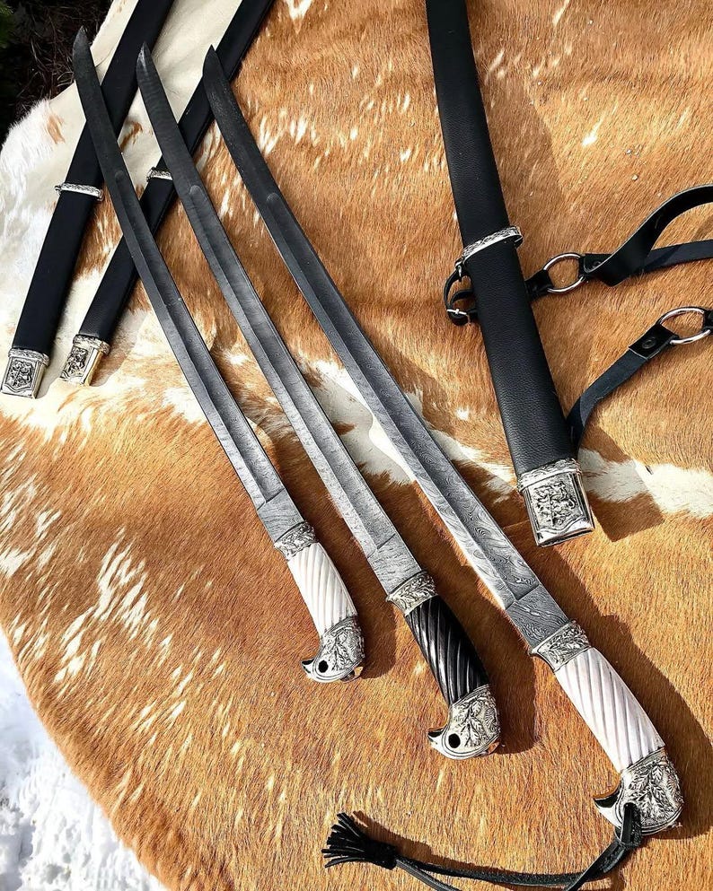 K&ouml;nnte beinhalten: Drei verzierte Schwerter mit aufwendigen Metallarbeiten und wei&szlig;en und schwarzen Griffen. Die Schwerter liegen auf einer braunen und wei&szlig;en Tierhaut.