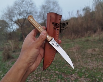 May include: A stainless steel knife with a wooden handle and a leather sheath. The knife has a curved blade and is held in front of a natural background. The text "MODEL KNIVES" is visible on the sheath.