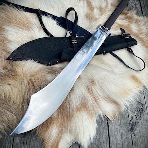 May include: A silver sword with a curved blade and a black leather scabbard with a strap. The sword is lying on a white and brown fur rug.