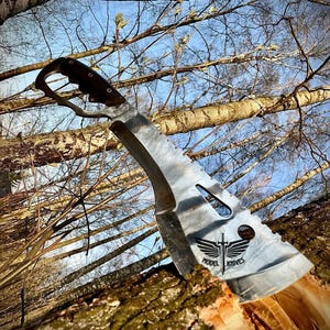 May include: A silver machete with a black handle and a black logo that says "Model Knives" with a sword and wings. The machete is stuck in a tree trunk.