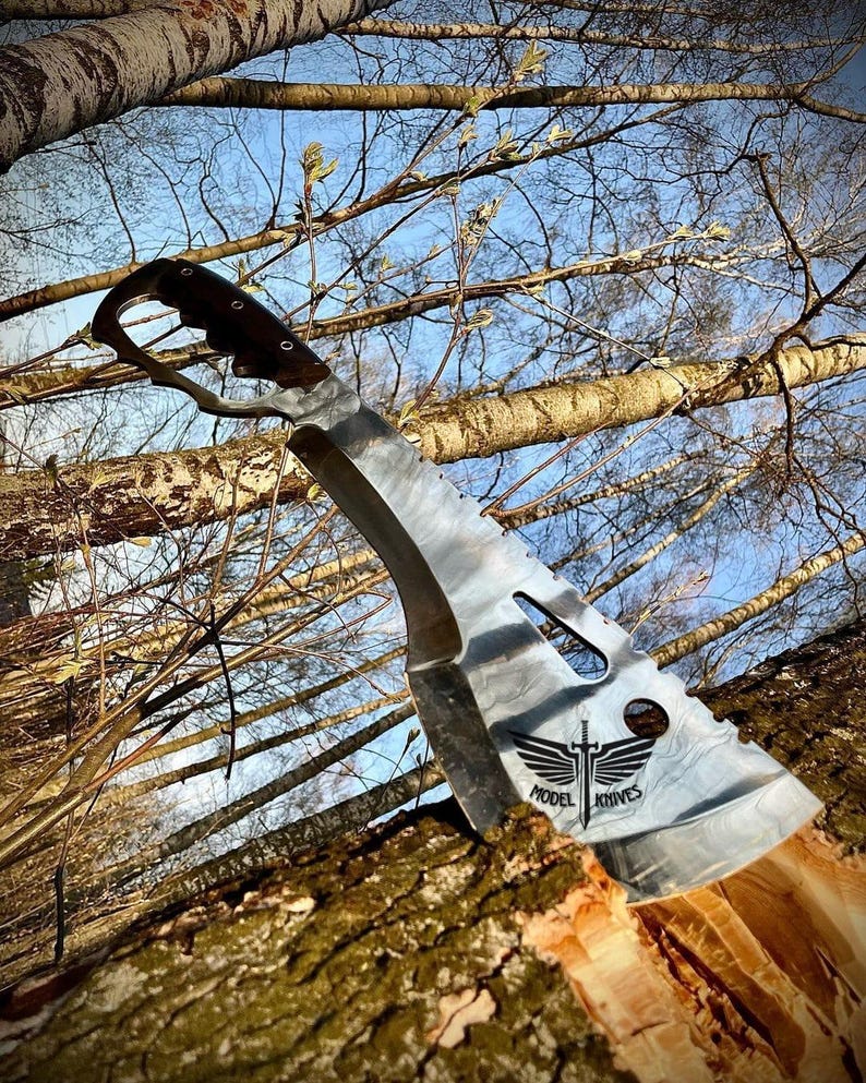 May include: A silver machete with a black handle and a black logo that says "Model Knives" with a sword and wings. The machete is stuck in a tree trunk.