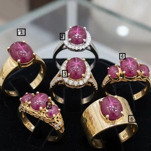 May include: A collection of six rings featuring oval-cut, star-effect rubies. The rings are set in gold and silver bands, with some adorned with surrounding diamonds. The ruby colors range from deep pink to red.