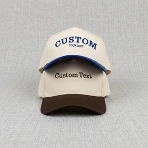 May include: Two custom baseball caps, one on top of the other. The top cap is cream-coloured with a blue brim and the word "CUSTOM" in blue script. The bottom cap is cream-coloured with a brown brim and the words "Custom Text".