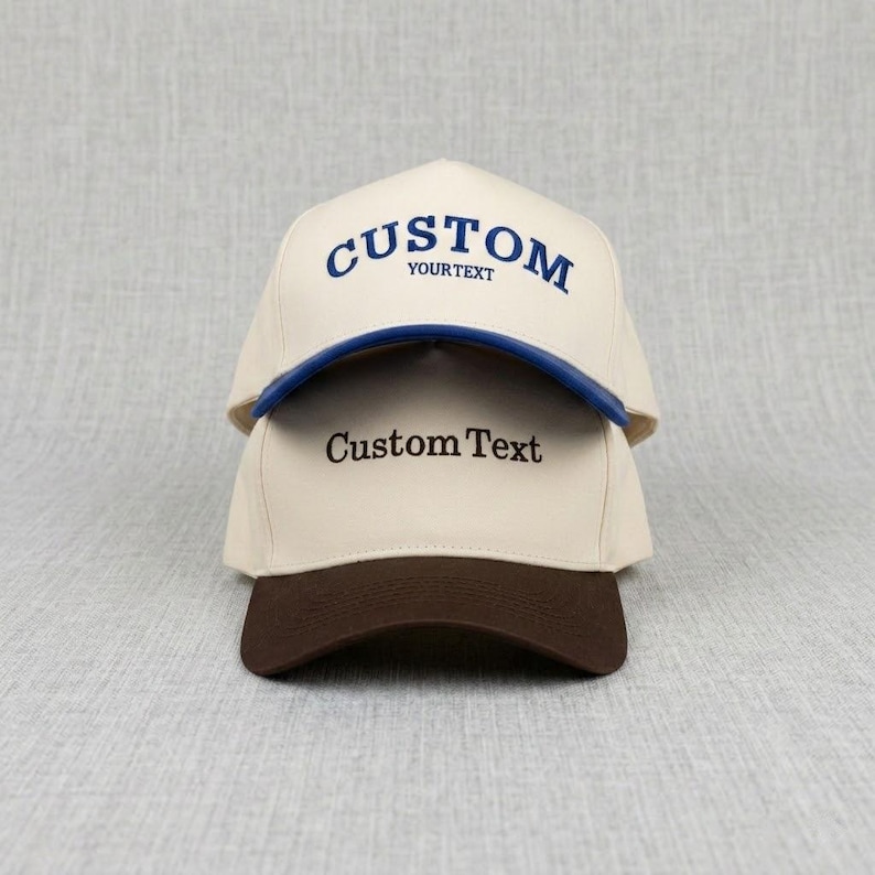 May include: Two custom baseball caps, one on top of the other. The top cap is cream-coloured with a blue brim and the word "CUSTOM" in blue script. The bottom cap is cream-coloured with a brown brim and the words "Custom Text".