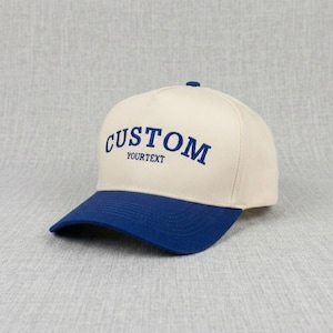 May include: A cream-coloured baseball cap with a blue brim. The word "CUSTOM" is embroidered in blue across the front, with "YOURTEXT" below it. The cap has a classic, structured design.