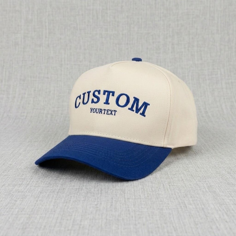 May include: A cream-coloured baseball cap with a blue brim. The word "CUSTOM" is embroidered in blue across the front, with "YOURTEXT" below it. The cap has a classic, structured design.