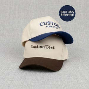 May include: Two custom baseball caps stacked on top of each other. The top cap is cream-colored with a navy blue brim and the text "CUSTOM YOUR TEXT". The bottom cap is cream-colored with a brown brim and the text "Custom Text".