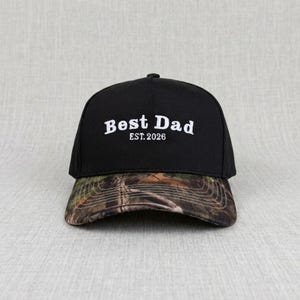 May include: Black baseball cap with a camouflage brim. The cap features the white text "Best Dad" above "EST. 2026". The hat is a classic six-panel design with a curved brim and adjustable strap.