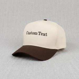 May include: A beige baseball cap with a brown peak. The cap has the embroidered text "Custom Text" in dark brown. It features a curved peak and a button on the crown.
