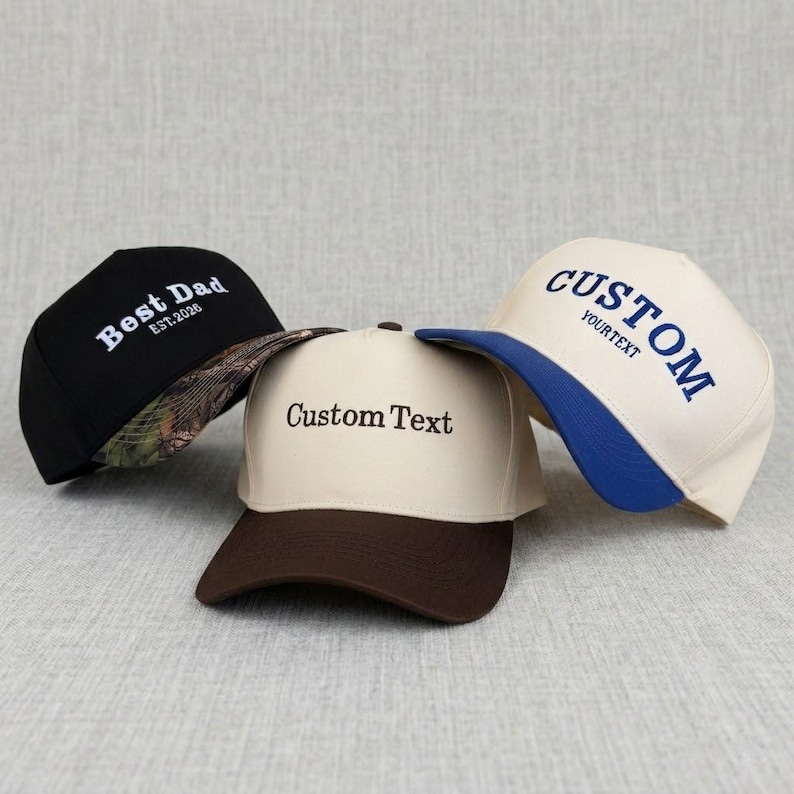 May include: Three baseball caps with custom text. One black cap has "Best Dad" embroidered in white, with a camouflage brim. A tan cap has "Custom Text" in black. A white cap has "CUSTOM YOUR TEXT" in blue.