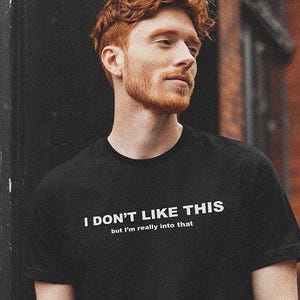 May include: A black short-sleeved t-shirt featuring the white text I DON'T LIKE THIS but I'm really into that printed across the chest. The shirt has a crew neckline and a standard fit, showcasing a clear graphic design on the front of the garment.