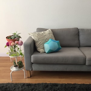 Op de afbeelding: Een grijze bank met twee decoratieve kussens. Eén kussen heeft een bladerpatroon, de andere een geometrische vorm in blauw- en groentinten. Een kleine witte plantenstandaard houdt bloemen en planten vast.