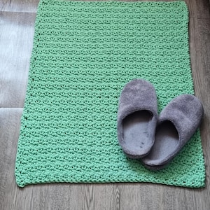 May include: A handmade, light green crochet bath mat with a textured surface. Two gray, plush slippers rest on the mat. The mat and slippers are on a wooden floor.