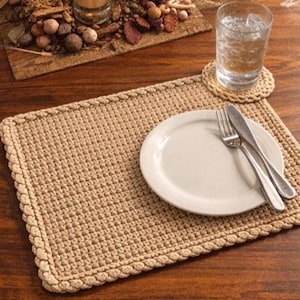 May include: A beige crocheted placemat with a decorative border, holding a white plate, fork, and knife. A glass of water sits on a matching coaster. The scene is set on a dark wooden table.