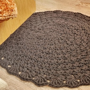May include: A circular dark brown crochet rug featuring a textured stitch pattern throughout the center and a decorative scalloped edge design. The rug is crafted from thick yarn and maintains a consistent dark brown color across its entire surface.