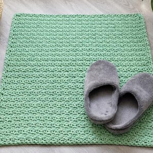 May include: A rectangular, light green crocheted rug with a textured, knotted stitch pattern. A pair of soft, grey, closed-toe slippers with a plush, fuzzy fabric finish are placed on the right side of the rug, resting on the woven surface.