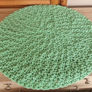 May include: A round, light green crocheted cushion. The cushion is made of thick yarn and has a textured surface. The item is sitting on a wooden surface.