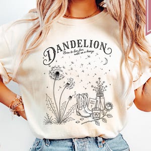 May include: Ivory-colored t-shirt with a black graphic of a dandelion, boots, and a Bible. The text reads "Dandelion, Born to live free, wild on a breeze." A pink banner reads "Super Soft."
