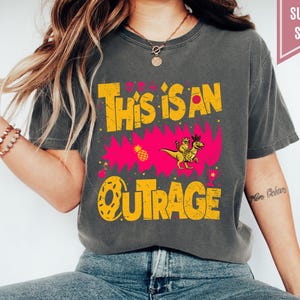 May include: Dark gray t-shirt with the text "THIS IS AN OUTRAGE" in yellow, featuring a cartoon dinosaur and a pink burst design. The shirt has a relaxed fit and a vintage look.