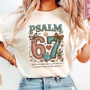 May include: Ivory-colored t-shirt with the text "PSALM 67" in teal and pink, featuring decorative elements like a star, heart, and cactus. The shirt also includes the quote "God be merciful unto us, and bless us; and cause his face to shine upon us."