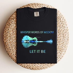 Puede incluir: Camiseta negra con el texto "WHISPER WORDS OF WISDOM" sobre un dise&ntilde;o en forma de guitarra con un &aacute;rbol y un reflejo de agua, y las palabras "LET IT BE" debajo. El dise&ntilde;o utiliza tonos azules y verdes.