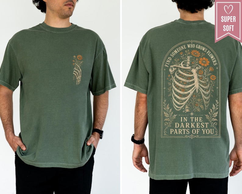 May include: An olive green t-shirt featuring a small skeletal hand holding flowers on the front left chest. The back displays a large graphic of a human ribcage intertwined with orange flowers and the text, Find someone who grows flowers in the darkest parts of you.