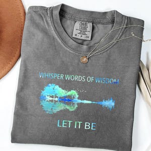 Puede incluir: Una camiseta gris Comfort Colors con el texto "Whisper Words of Wisdom" y "Let It Be" en degradado azul-verde. Un gr&aacute;fico de guitarra est&aacute; en el centro. Un collar plateado con un colgante est&aacute; en la camisa.