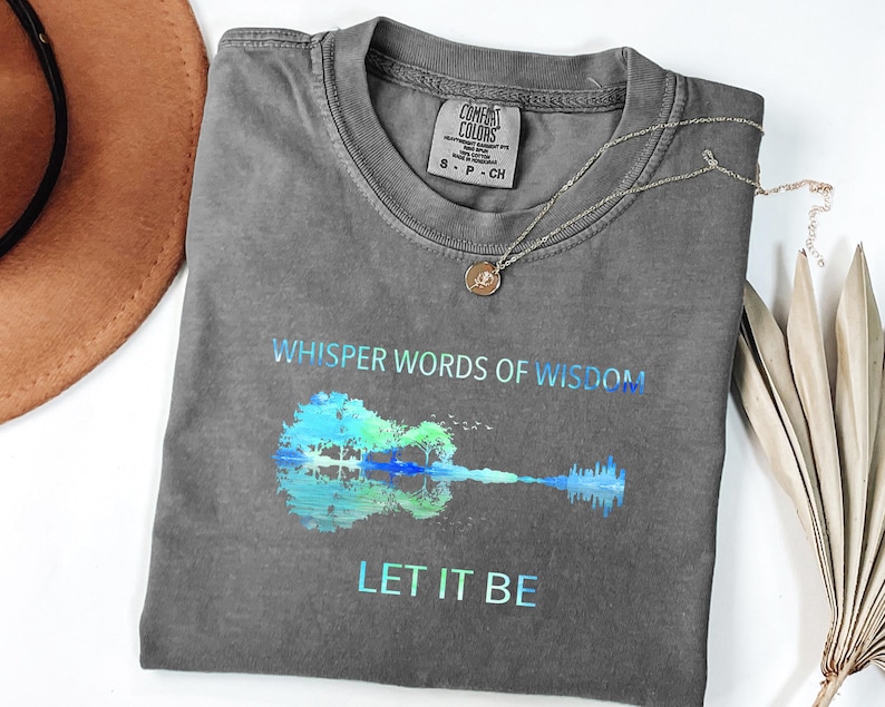 Puede incluir: Una camiseta gris Comfort Colors con el texto "Whisper Words of Wisdom" y "Let It Be" en degradado azul-verde. Un gr&aacute;fico de guitarra est&aacute; en el centro. Un collar plateado con un colgante est&aacute; en la camisa.