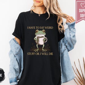 May include: Black t-shirt featuring a cartoon frog holding a coffee cup. The frog is green and white, and the shirt has the text "I HAVE TO SAY WEIRD STUFF OR I WILL DIE" in a serif font. A pink banner reads "SUPER SOFT".