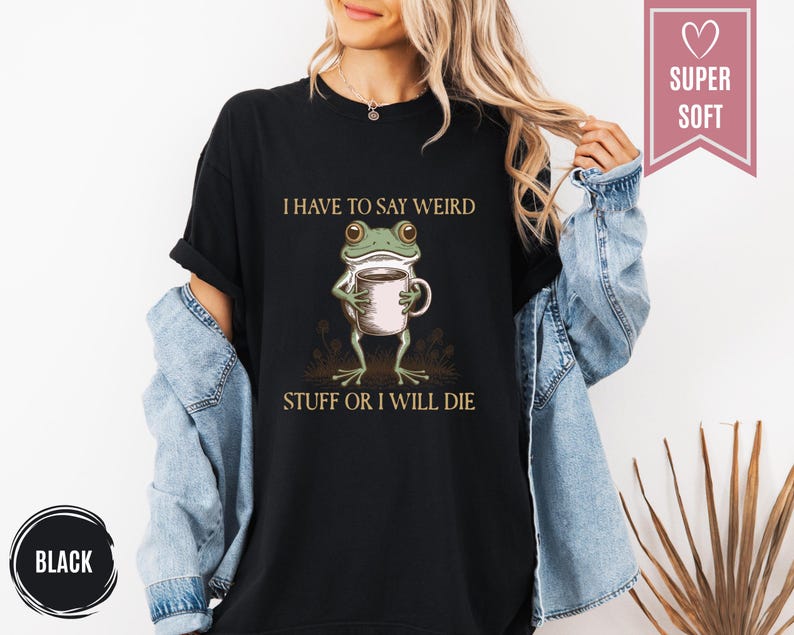 May include: Black t-shirt featuring a cartoon frog holding a coffee cup. The frog is green and white, and the shirt has the text "I HAVE TO SAY WEIRD STUFF OR I WILL DIE" in a serif font. A pink banner reads "SUPER SOFT".