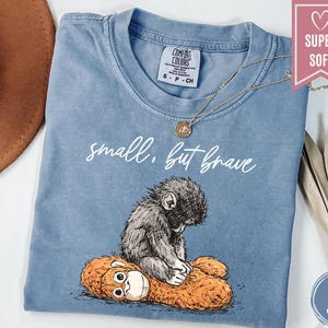 May include: A white ceramic mug with a black handle and the text "But First, Coffee" in a bold, black font. The mug is set against a blurred background, suggesting a cozy setting.