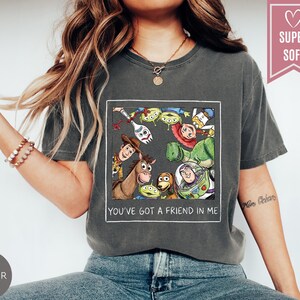 Op de afbeelding: Donkergrijs T-shirt met een kleurrijke afbeelding van personages uit de animatiefilm Toy Story. De afbeelding bevat Woody, Buzz Lightyear en andere personages. De tekst "YOU'VE GOT A FRIEND IN ME" staat eronder.