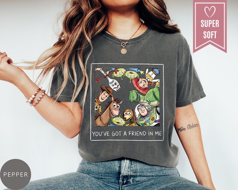 Op de afbeelding: Donkergrijs T-shirt met een kleurrijke afbeelding van personages uit de animatiefilm Toy Story. De afbeelding bevat Woody, Buzz Lightyear en andere personages. De tekst "YOU'VE GOT A FRIEND IN ME" staat eronder.