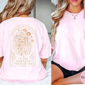 May include: A light pink t-shirt featuring a graphic on the back of a human rib cage intertwined with orange and gold flowers, with the text "FIND SOMEONE WHO GROWS FLOWERS IN THE DARKEST PARTS OF YOU." A smaller matching floral rib cage graphic is on the front.