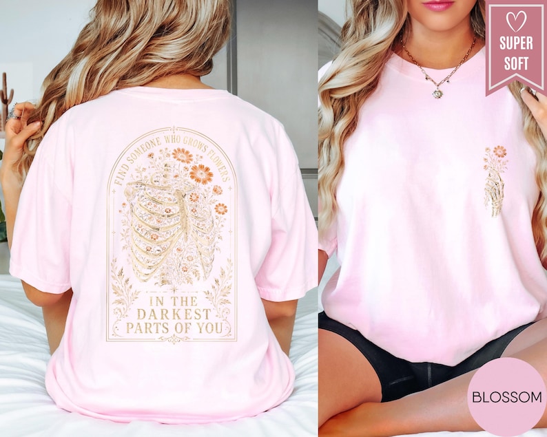 May include: A light pink t-shirt featuring a graphic on the back of a human rib cage intertwined with orange and gold flowers, with the text "FIND SOMEONE WHO GROWS FLOWERS IN THE DARKEST PARTS OF YOU." A smaller matching floral rib cage graphic is on the front.