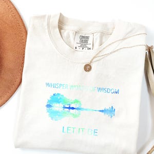 Puede incluir: Camiseta color crema con un dise&ntilde;o de guitarra en acuarela en tonos azules y verdes. Los textos "Whisper Words of Wisdom" y "Let It Be" est&aacute;n impresos encima y debajo de la guitarra. Tambi&eacute;n hay un collar dorado y un sombrero marr&oacute;n.