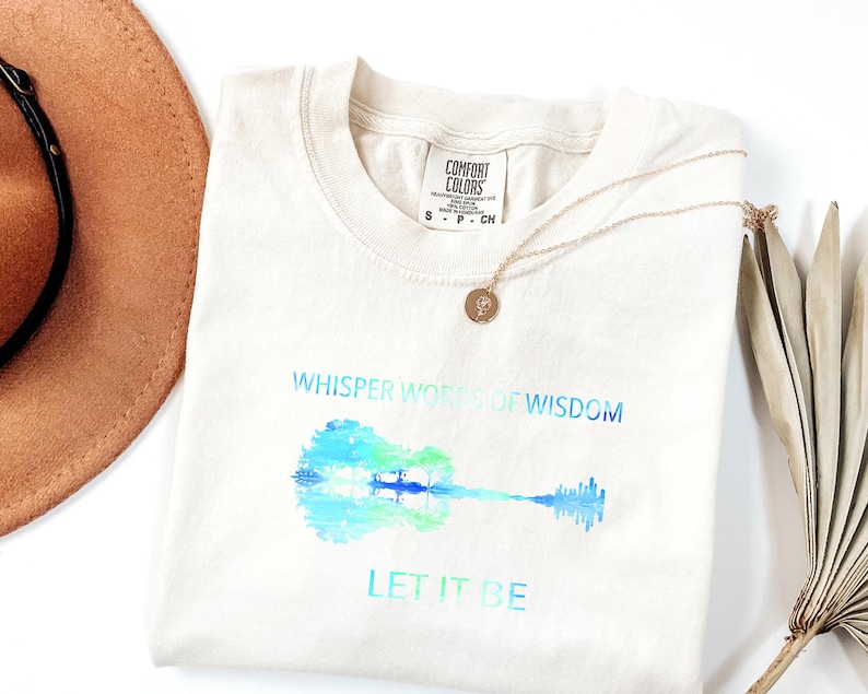 Puede incluir: Camiseta color crema con un dise&ntilde;o de guitarra en acuarela en tonos azules y verdes. Los textos "Whisper Words of Wisdom" y "Let It Be" est&aacute;n impresos encima y debajo de la guitarra. Tambi&eacute;n hay un collar dorado y un sombrero marr&oacute;n.
